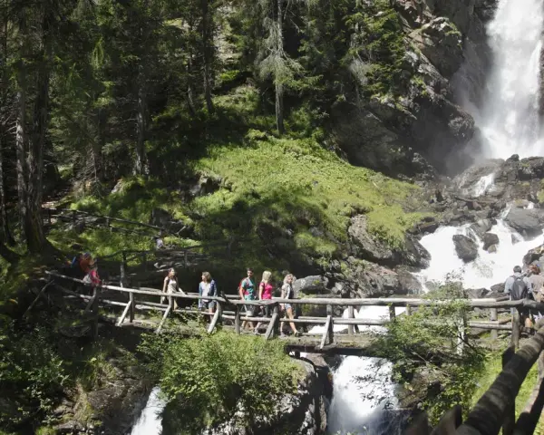 Gruppo di escursionisti attraversa un ponte di legno sul sentiero del Val di Rabbi, con una cascata impetuosa sullo sfondo.