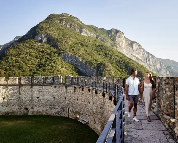 Coppia passeggia su un bastione di un castello con montagne verdi sullo sfondo. Un'esperienza da vivere nel Trentino da scoprire.