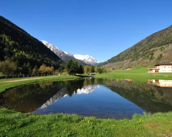 I laghetti di Vermiglio riflettono montagne innevate e un cielo azzurro. Un edificio moderno si specchia nell'acqua calma.