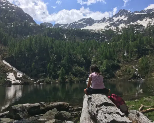 Bambina seduta su un tronco guarda il lago di Barco, circondato da montagne innevate e foreste di conifere.