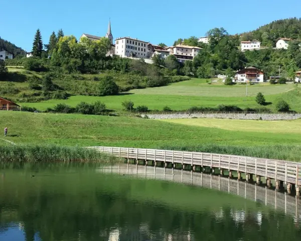 Una piacevole passeggiata dria al fos, con un ponte di legno che attraversa il lago e un villaggio sullo sfondo.