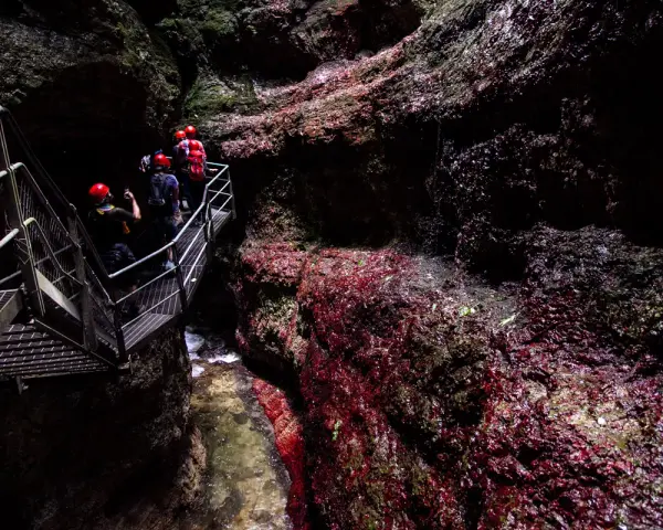 Esploratori con caschi rossi percorrono una passerella metallica nel **canyon Rio Sass fondo**, tra rocce scure e rosse.