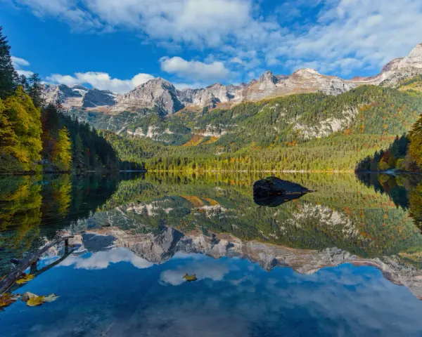 Il **lago di Tovel** riflette montagne maestose e boschi autunnali sotto un cielo azzurro con nuvole bianche. Una roccia emerge dall'acqua cristallina.