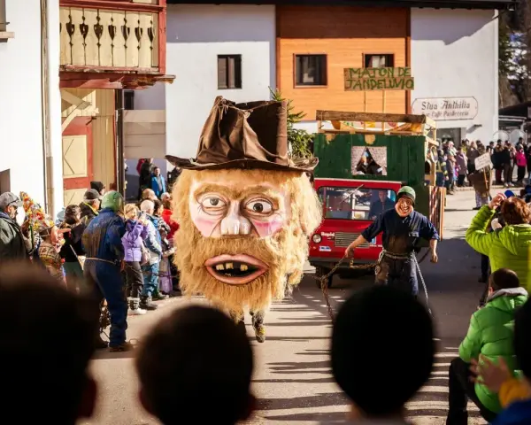 Gigantesca maschera pelosa con cappello marrone sfila al carnevale trentino. Un uomo con maschera grottesca trascina una catena.
