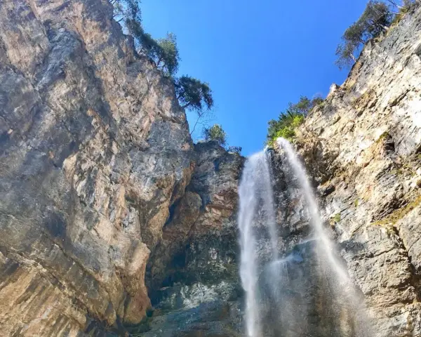 La cascata di Tret scende maestosa da una parete rocciosa imponente, sotto un cielo azzurro e limpido.