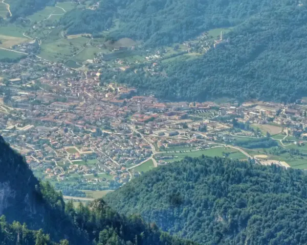 Vista aerea della Valsugana, Trento, con un paese incastonato tra montagne verdi e un picco roccioso in primo piano.