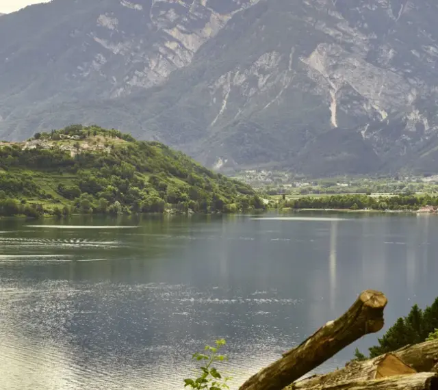 Villaggio su collina verde affacciato sul lago di Caldonazzo, con montagne imponenti sullo sfondo.