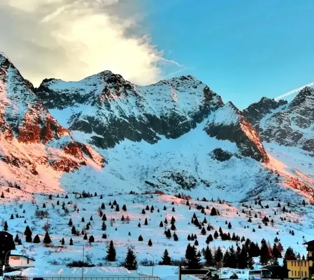 Montagne innevate al tramonto sul Passo del Tonale, con alberi sparsi e un hotel.
