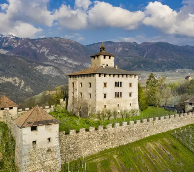Castel Nanno, un maniero fortificato con torri e mura di pietra, domina un paesaggio collinare con vigneti e montagne sullo sfondo.