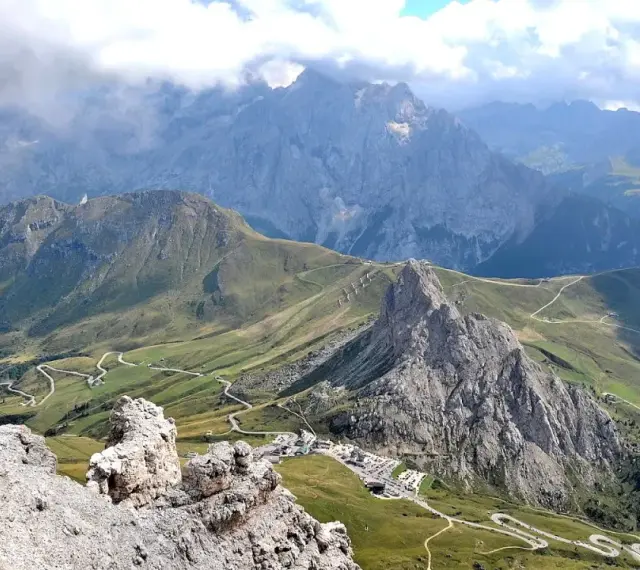Panoramica mozzafiato del **Passo Pordoi** con strade tortuose che si snodano tra verdi pendii e imponenti vette rocciose, sotto un cielo nuvoloso.
