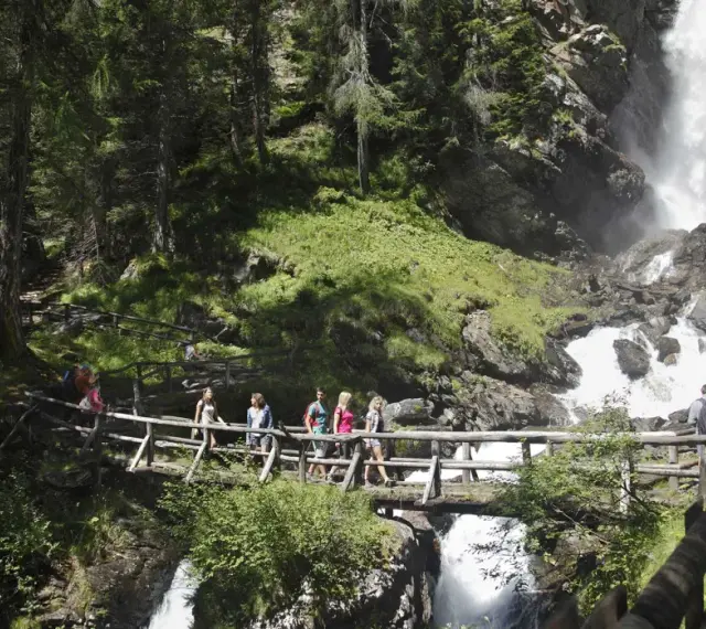 Gruppo di escursionisti attraversa un ponte di legno sul sentiero del Val di Rabbi, con una cascata impetuosa sullo sfondo.