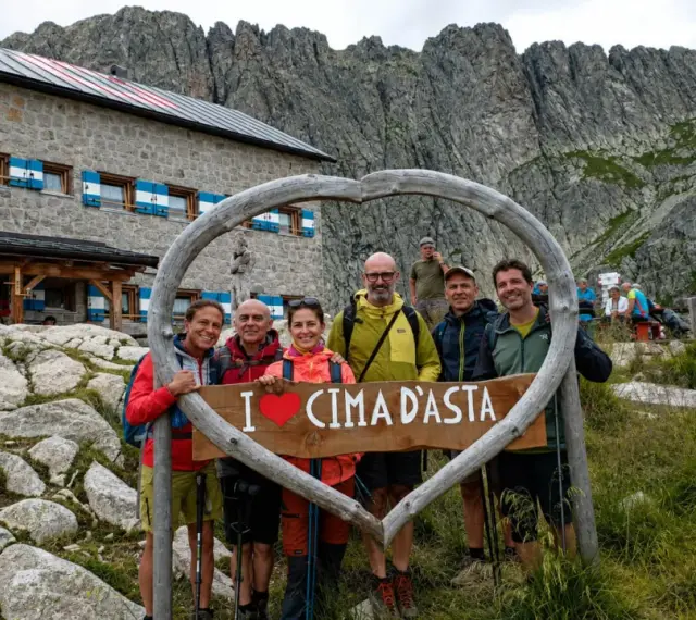 Gruppo di escursionisti felici davanti a un rifugio alpino, con un cartello a forma di cuore che dice "I ❤️ CIMA D'ASTA". Un'indimenticabile esperienza di trekking Trentino.