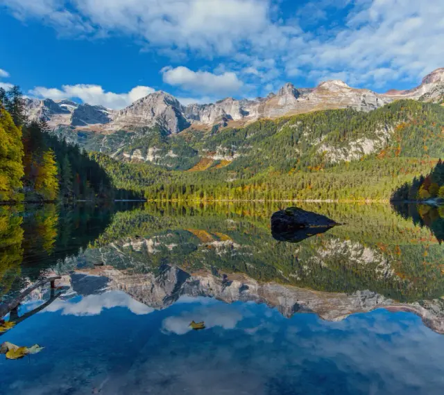 Il **lago di Tovel** riflette montagne maestose e boschi autunnali sotto un cielo azzurro con nuvole bianche. Una roccia emerge dall'acqua cristallina.