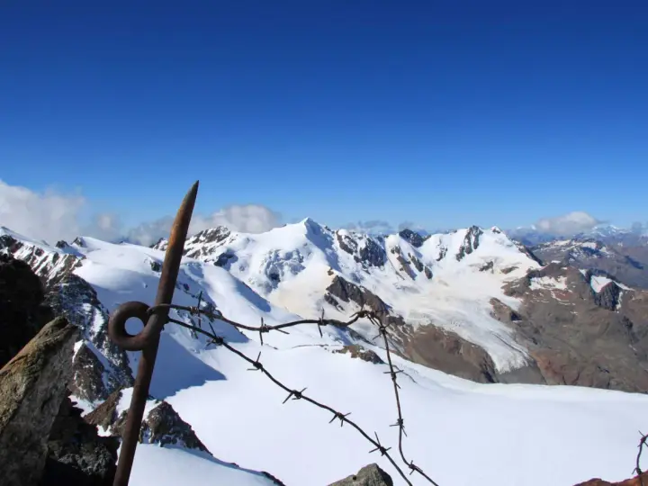 Vista mozzafiato dalla punta Linke, con cime innevate e un cielo azzurro intenso. Un vecchio piccone arrugginito e filo spinato segnano il confine.
