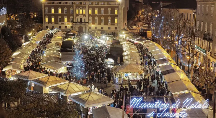 Un affollato Mercatino di Natale a Trento, con bancarelle illuminate, un albero scintillante e gente che passeggia tra le luci festive.