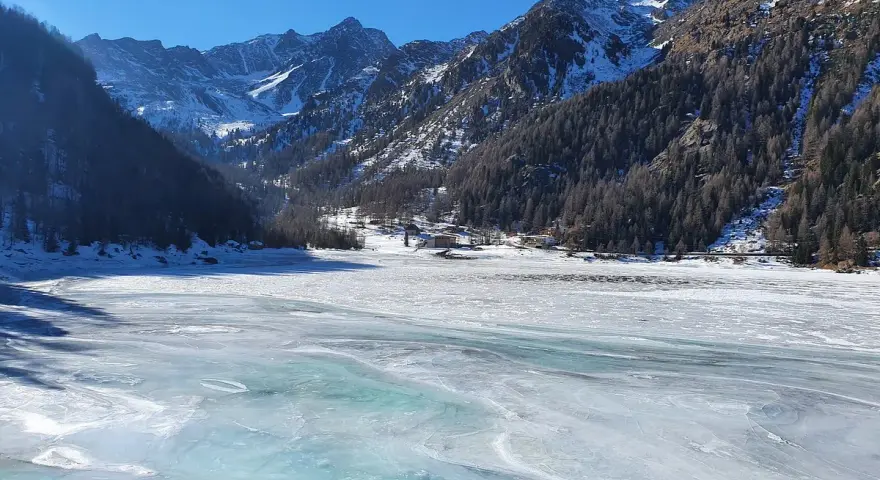 Il lago di Fontana Bianca, un'immensa distesa ghiacciata sotto un cielo azzurro, circondato da montagne innevate e boschi scuri.