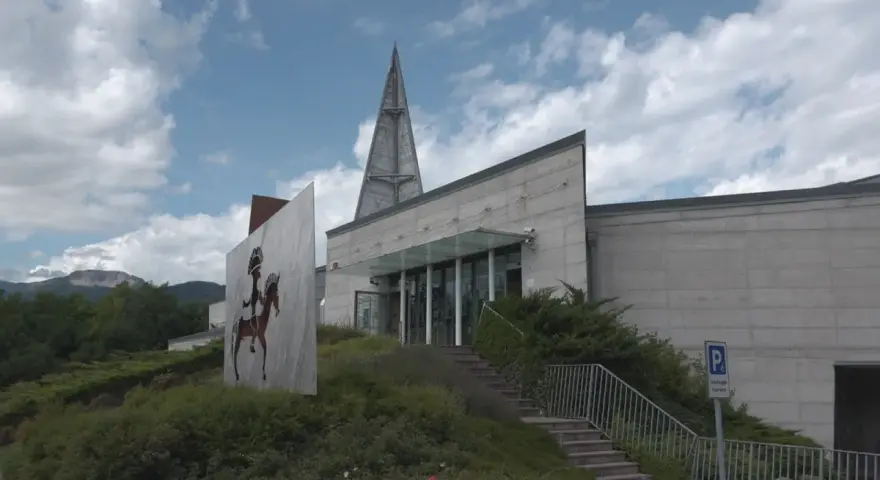 Il museo retico, con la sua architettura moderna e un'installazione artistica di un guerriero a cavallo, si erge sotto un cielo nuvoloso.