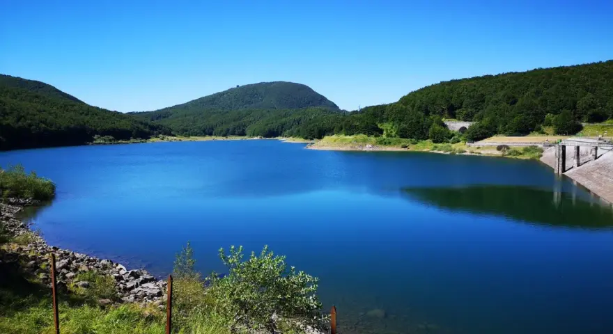 Vasto lago di Comano con acque blu intenso, circondato da colline verdi e una diga in cemento.