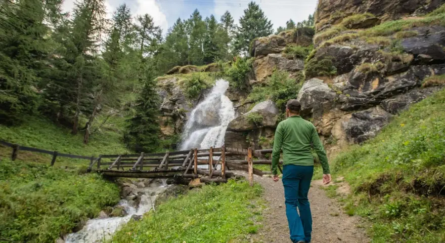 Un escursionista ammira una cascata nel Val di Sole in estate, con un ponte di legno che attraversa il torrente.