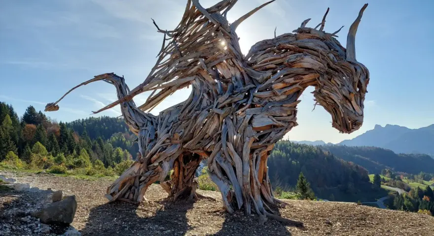 Un maestoso drago di Vaia, scolpito nel legno, domina un paesaggio montano con le ali spiegate verso il sole.