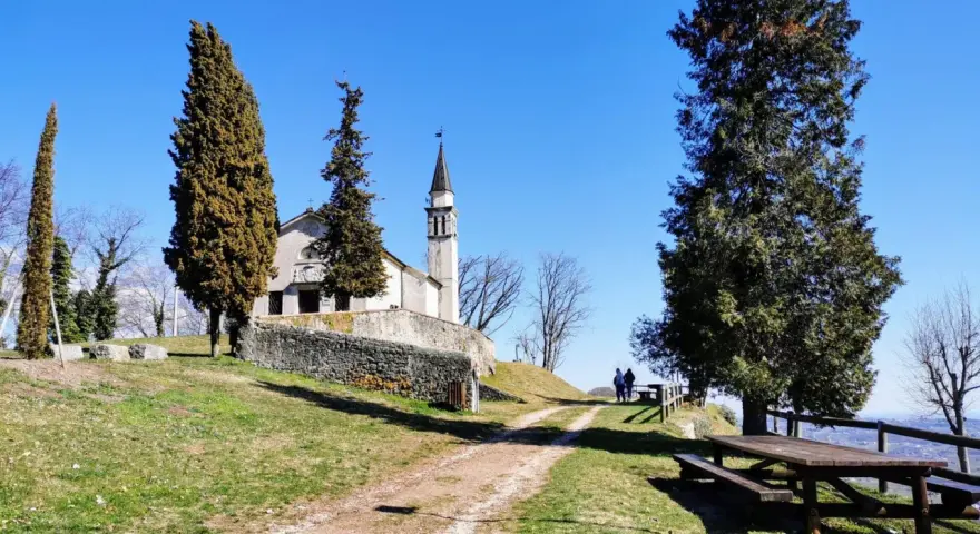 A church with a bell tower sits on a grassy hill, with tall trees and a picnic table in the foreground.