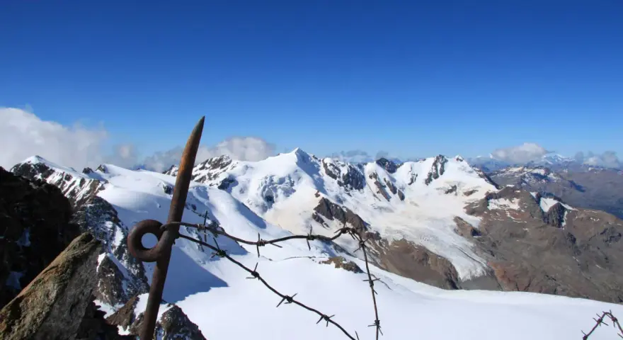 Vista mozzafiato dalla punta Linke, con cime innevate e un cielo azzurro intenso. Un vecchio piccone arrugginito e filo spinato segnano il confine.