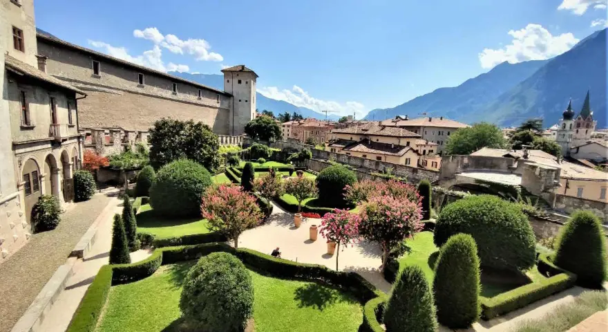 Giardini curati e fioriti incorniciano il maestoso Castel Buonconsiglio, con torri imponenti e montagne sullo sfondo.