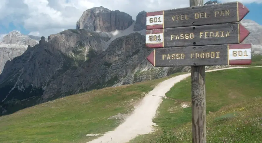 Cartello con indicazioni per Viel del Pan, Passo Fedaia e Passo Pordoi. Sentiero in salita tra prati verdi e montagne imponenti.