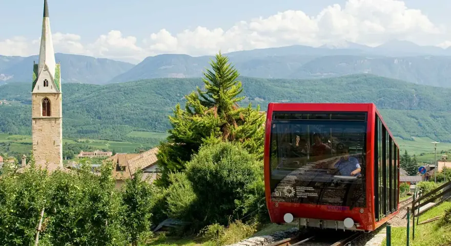 Funicolare rossa in Trentino Alto Adige, con campanile e montagne sullo sfondo. Un'ottima idea per scoprire cosa vedere!