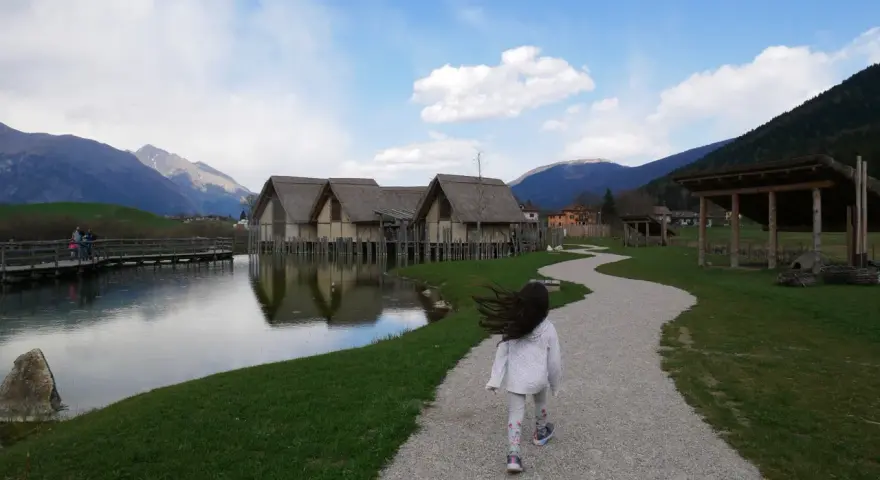 Bambina corre su sentiero ghiaioso verso villaggio palafitticolo nelle Alpi Giudicarie. Montagne sullo sfondo.