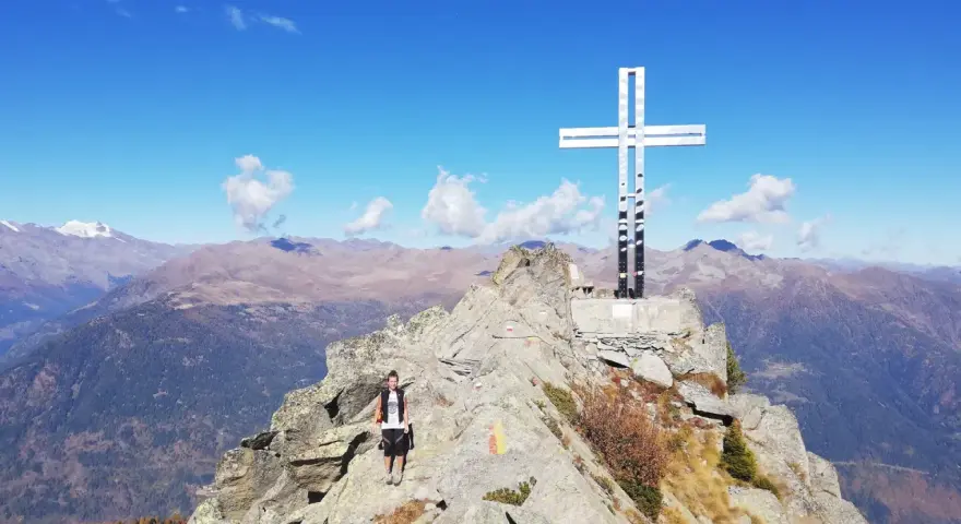Escursionista raggiunge la vetta con la croce della pace, circondata da montagne maestose sotto un cielo azzurro.