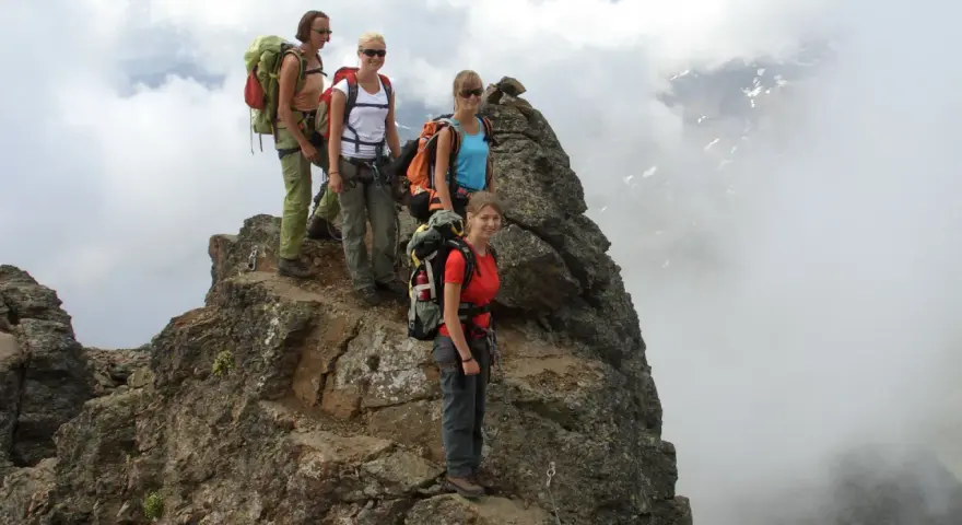 Quattro escursionisti con zaini si fermano sulla cima rocciosa di Cima Sternai, circondati da nuvole e nebbia.