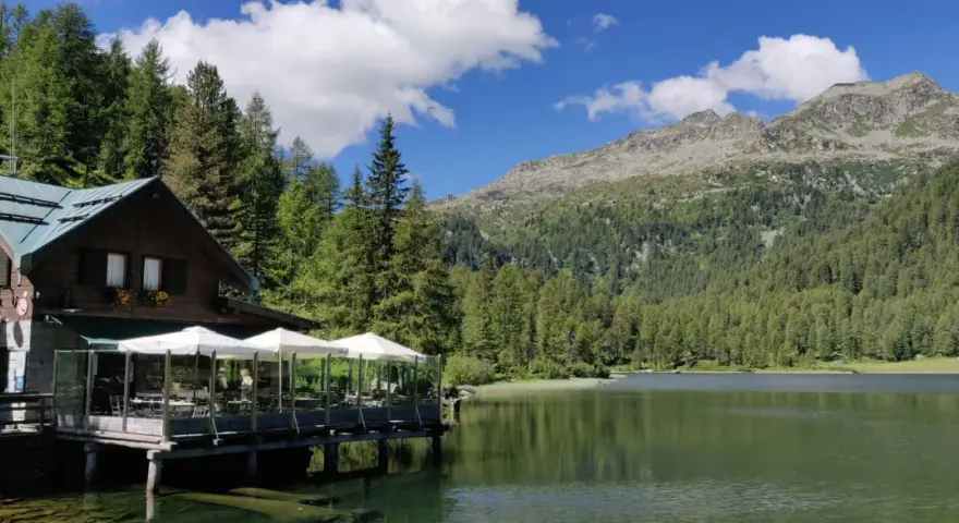 Rifugio in legno con tavoli all'aperto sul lago, circondato da boschi e montagne. Un luogo incantevole per scoprire cosa vedere a Pellizzano.