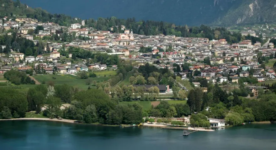 Vista panoramica del **lago di Levico** con il paese arroccato sulla collina. Alberi verdi e case colorate si specchiano nell'acqua tranquilla.