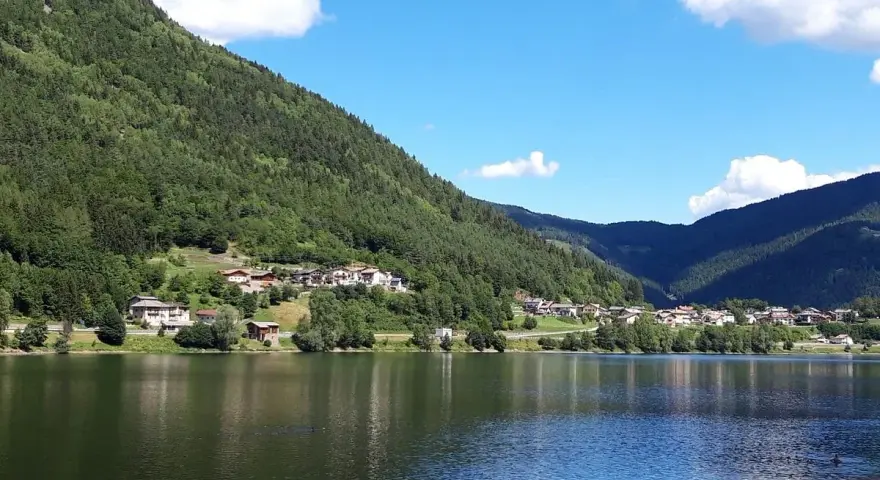 Il **lago di Serraia** riflette le montagne boscose e un villaggio alpino sotto un cielo azzurro con nuvole bianche.