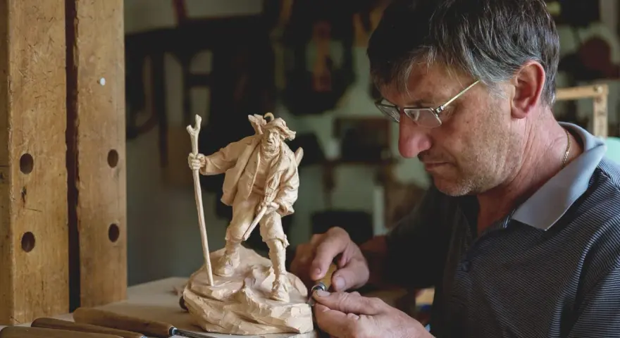 Maestro intaglia un pastore con bastone, un esempio di artigianato legno trentino.