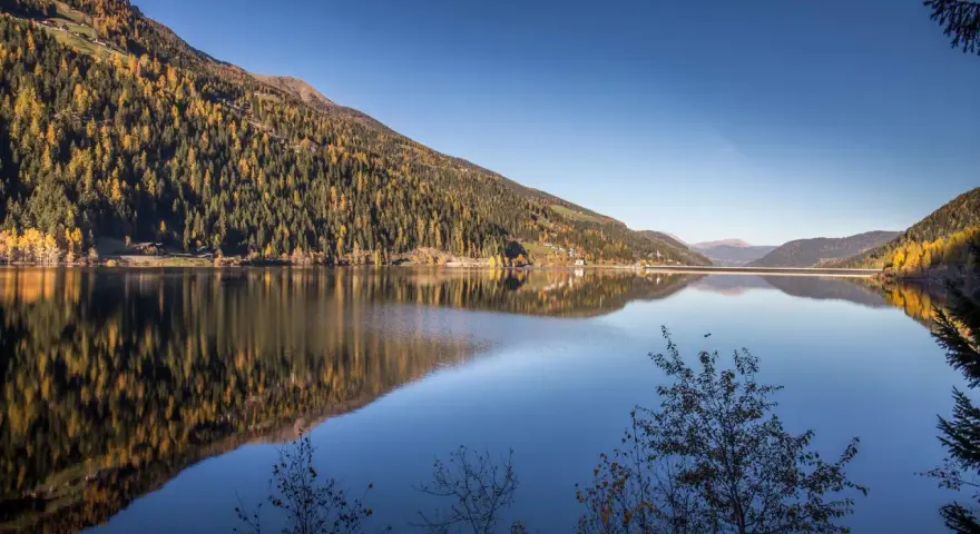 Autunno dorato sul **Val d'Ultimo lago**, con boschi che si specchiano nelle acque calme e un cielo azzurro intenso.
