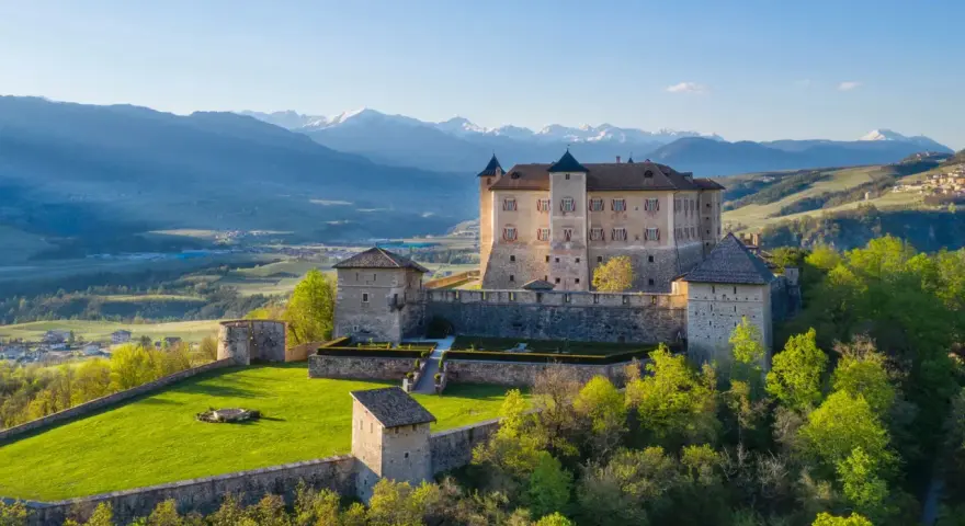 Castelli Val di Non, un imponente maniero su una collina verdeggiante, con montagne innevate sullo sfondo.