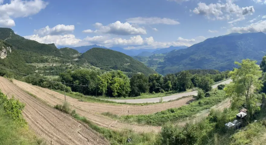 Panoramica del passo di Santa Barbara a Trento, con campi coltivati, alberi verdi e montagne sullo sfondo.