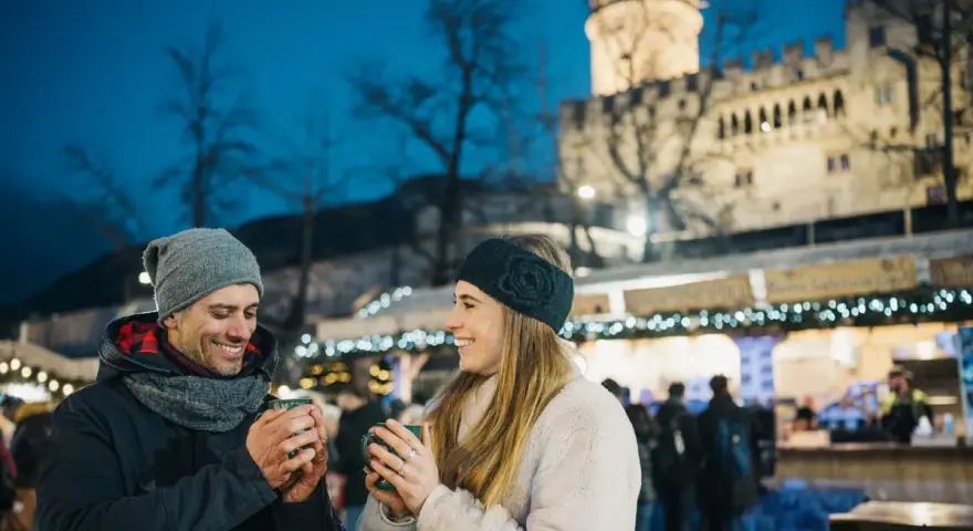 Coppia sorridente con tazze fumanti al mercatino di Natale di Trento inverno, con il castello illuminato sullo sfondo.