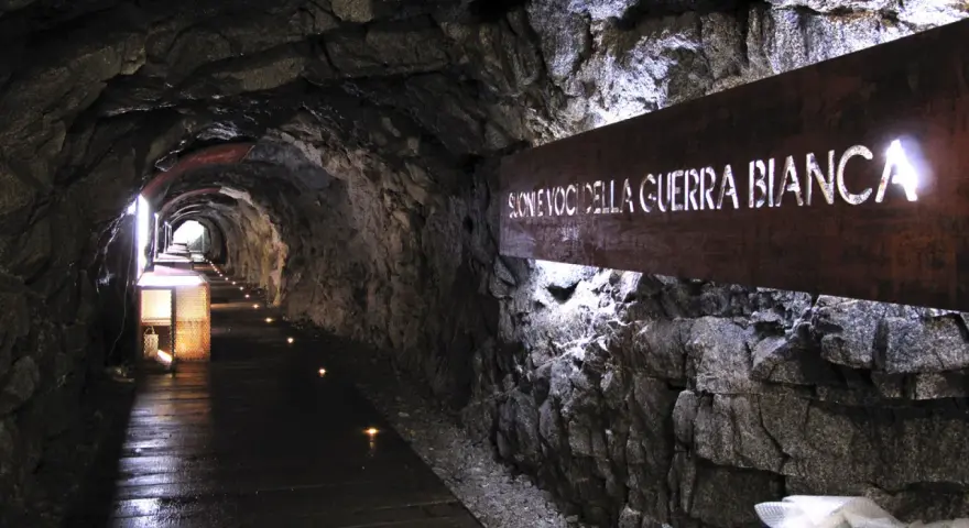 Un suggestivo tunnel roccioso illuminato, con un cartello che recita "Suoni voci della Guerra Bianca". Un percorso che sembra condurre alla galleria paradiso.