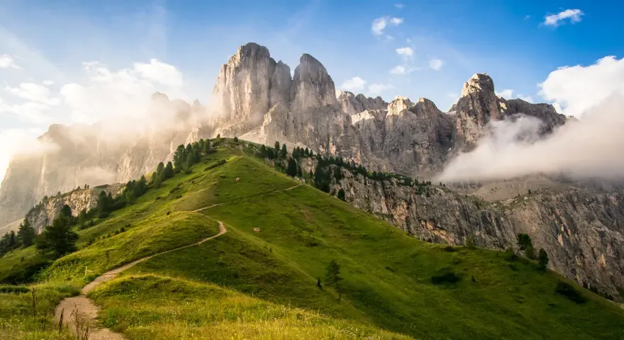 Sentiero serpeggiante su un prato verdeggiante ai piedi di un imponente gruppo montuoso tra Veneto e Trentino Alto Adige, con nuvole che accarezzano le cime rocciose.
