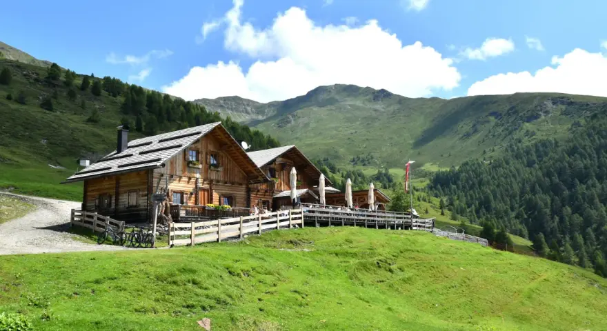 Rifugio in legno con tavoli all'aperto, ideale per un giro delle malghe in Val di Non. Montagne verdi e cielo azzurro.