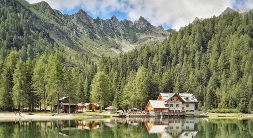 Riflessi sul lago alpino nella piana di Nambino, con boschi di conifere e montagne sullo sfondo.