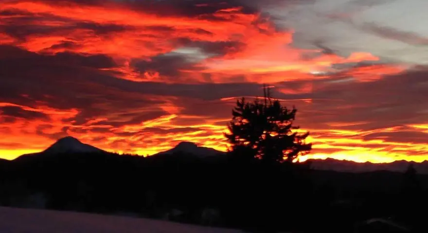 Un infuocato tramonto sulle Dolomiti, con nuvole rosse e arancioni che illuminano il cielo sopra le sagome scure delle montagne e di un pino solitario.