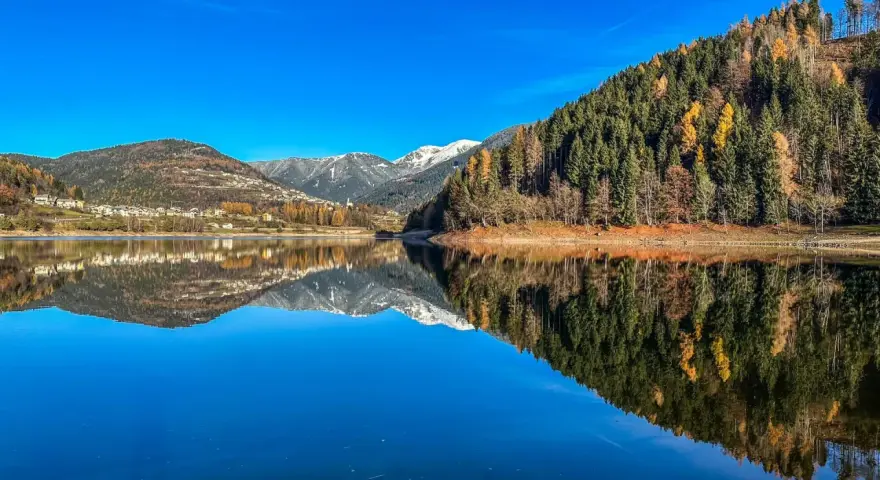Il lago delle Piazze riflette montagne innevate, boschi autunnali e un villaggio sotto un cielo azzurro.