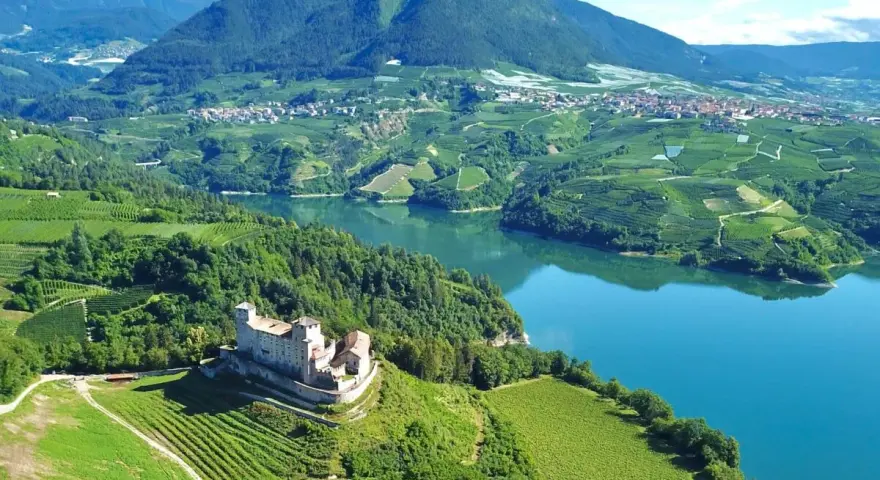 Castello medievale su una collina verdeggiante, con vigneti a terrazze e un lago azzurro nel **Val di Non**.