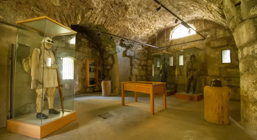 Interno di un museo in pietra con manichini in uniformi storiche, uno dei quali indossa un abito chiaro e porta un fucile, evocando un forte strino di battaglia.