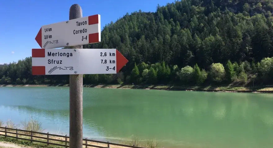 Cartello con indicazioni per Tavon di Coredo, Merlonga e Sfruz, con lago verde e bosco sullo sfondo.