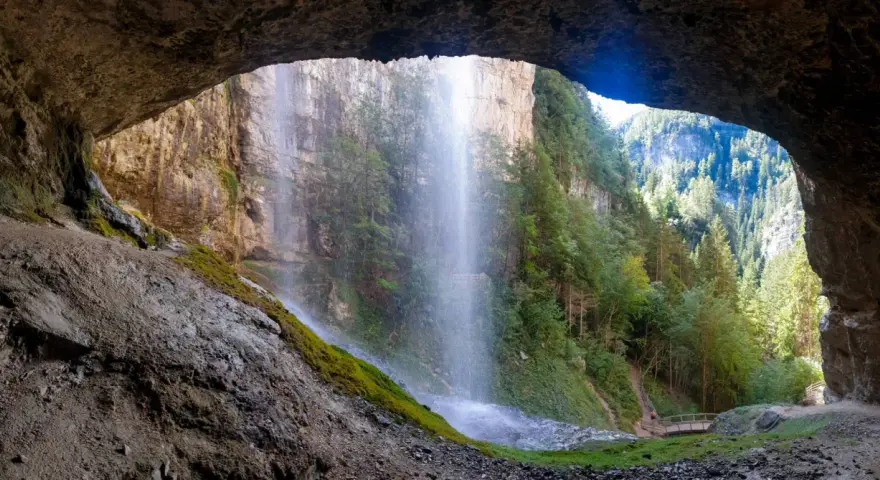 Vista da una grotta su maestose cascate del Trentino, con sentieri e ponticelli che invitano all'esplorazione.